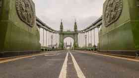 Hammersmith Bridge on a cloudy day. The picture is taken from the floor, and the lines of the road are at the centre of the image. The bridge surrounds the viewer. 