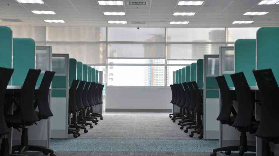 The picture shows a row of empty chairs and desks in an office. It is a well lit room filled with plenty of natural light. 